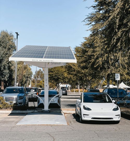 A white electric car charging under a solar panel in an urban setting, showcasing renewable energy use.